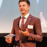 Porträt von Kevin.. Er steht auf einer Bühne und hält einen Vortrag. Er ist von Kopf bis zur Hüfte abgebildet. Kevin trägt einen rot-braunen eriösen Anzug, ein weißes emd und eine rote Krawatte. In seiner Hand hält er Moderationskarten. Im Hintergrund ist eine Leinwand zu sehen, auf der eine Präsentation läuft.