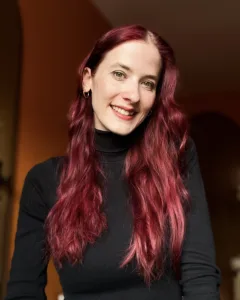 Porträt von Alina, einer jungen weißen Frau mit langen roten Locken. Sie trägt einrn schwarzen Pullover