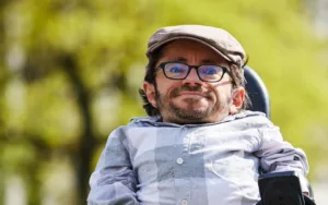 Porträt von Raúl, einem weißen Mann mit Peruanischer Herkunft. Er ist ein kleiner Mann mit hellbraunen Haaren, einem braunen Bart und einer Brille mit dickem, schwarzen Rand. Er ist draußen und trägt eine beige Mütze und ein hellblaues Hemd.